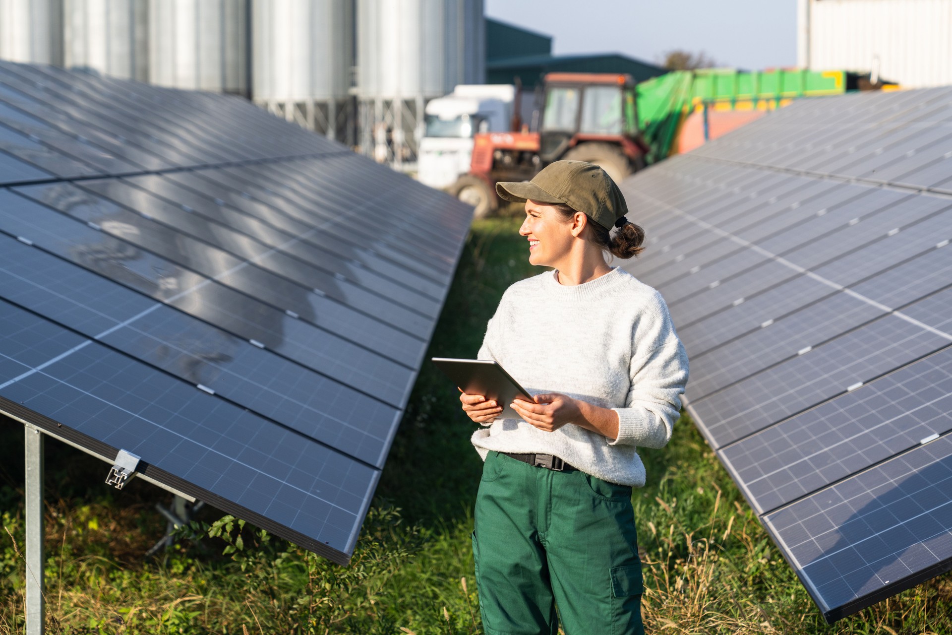 Female farmer wearing work uniform holds digital tablet in her hands stands next to solar panels. Modern agricultural farm using solar panels. Agricultural silos in the background.
